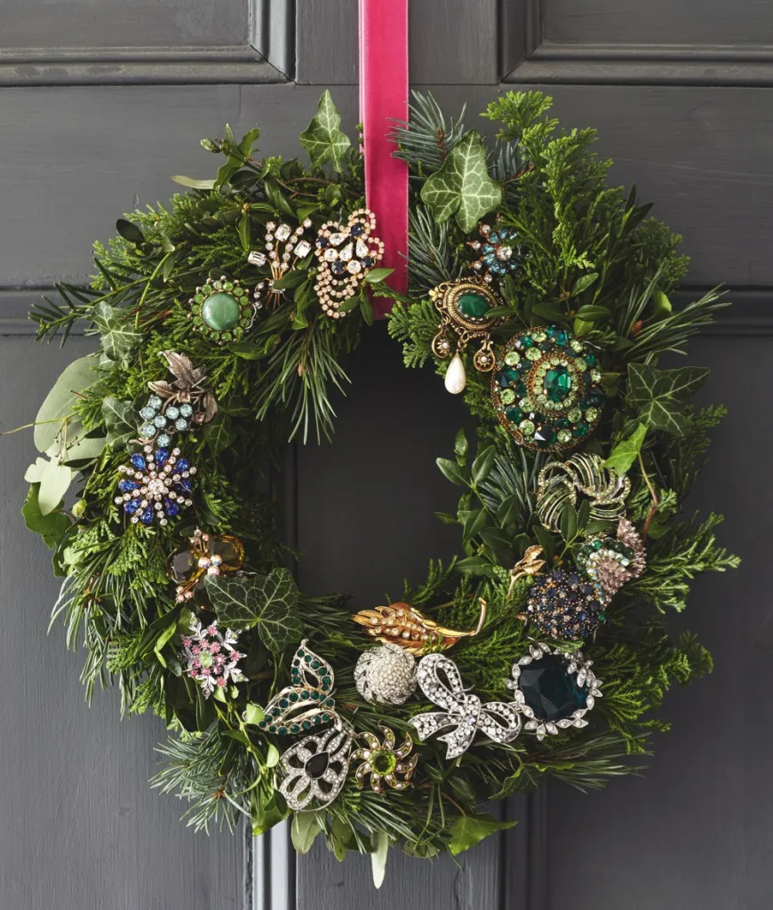A festive wreath hangs on a dark grey front door. The wreath is peppered with sparkling antique and vintage brooches.
