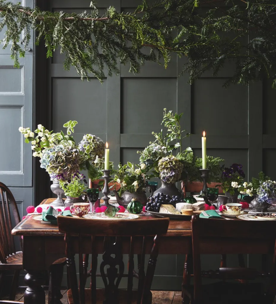 A Christmassy dining table in a dark grey living room. The table is decorated with antique pewter jugs filled with evergreen fronds and mophead hydrangeas. A string of pink neon pompoms brings some colour.