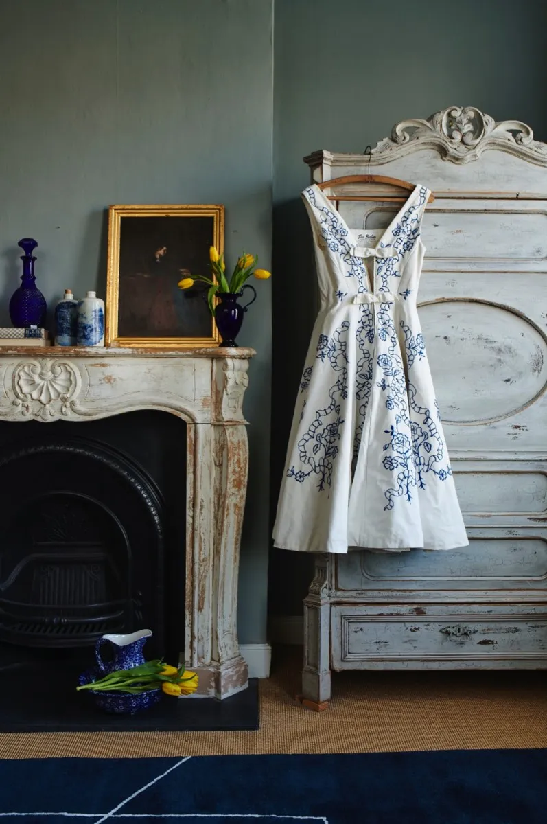 A white dress with a blue pattern on it hangs by a mantelpiece with a number of blue and white ceramics on it