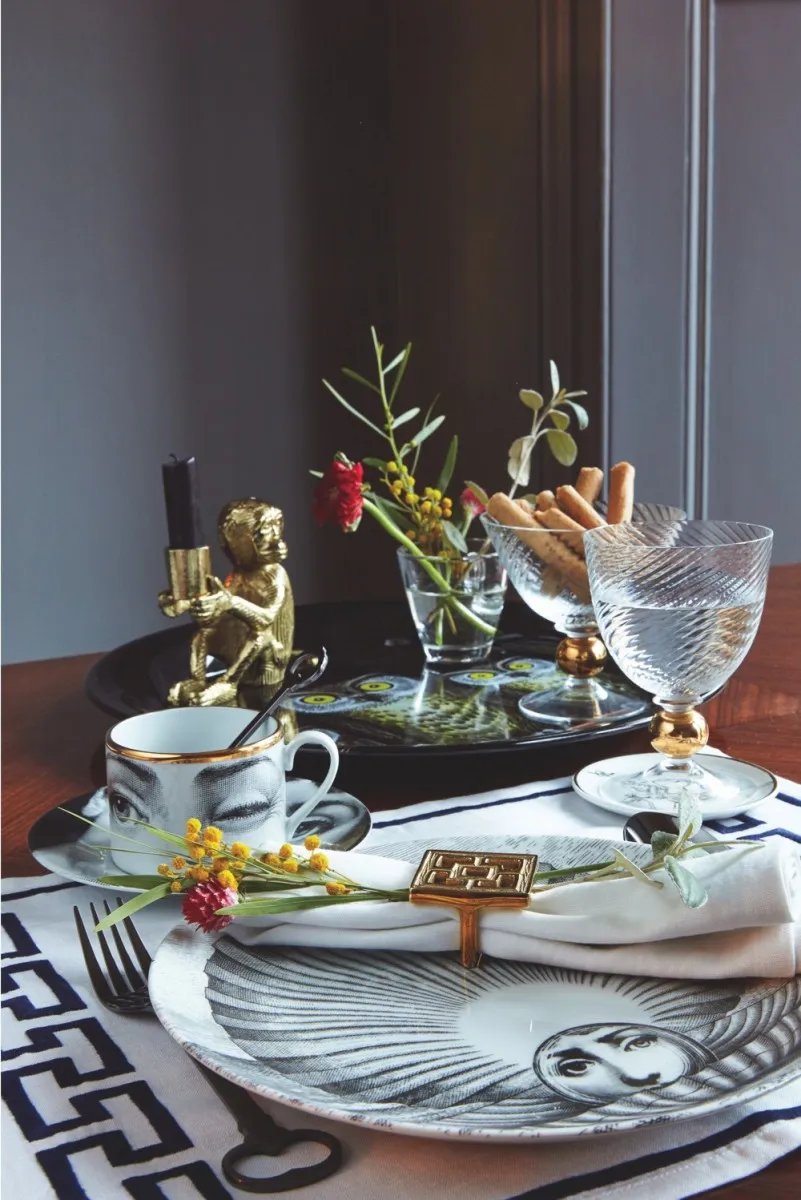 A brown breakfast table featuring a Fornasetti tray and tea cup, plus glasses filled with flowers and breadsticks