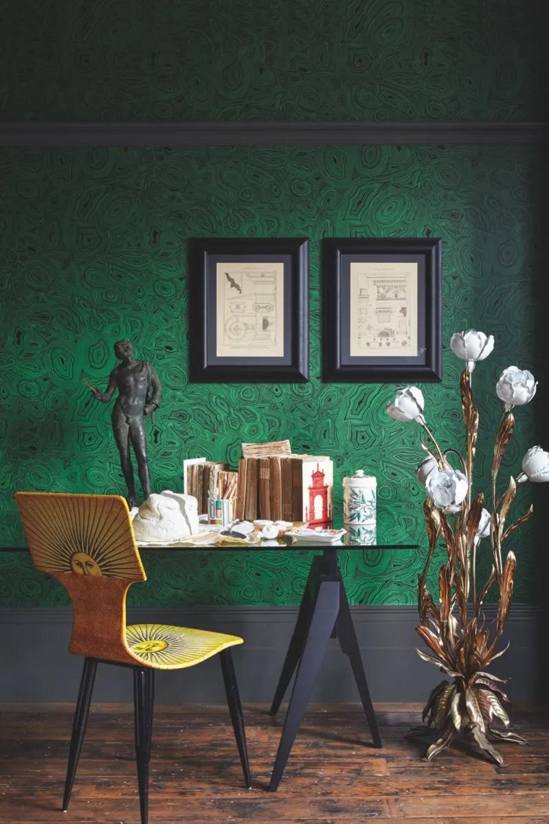 A yellow Fornasetti dining chair at a glass-topped writing desk against green malachite wallpaper