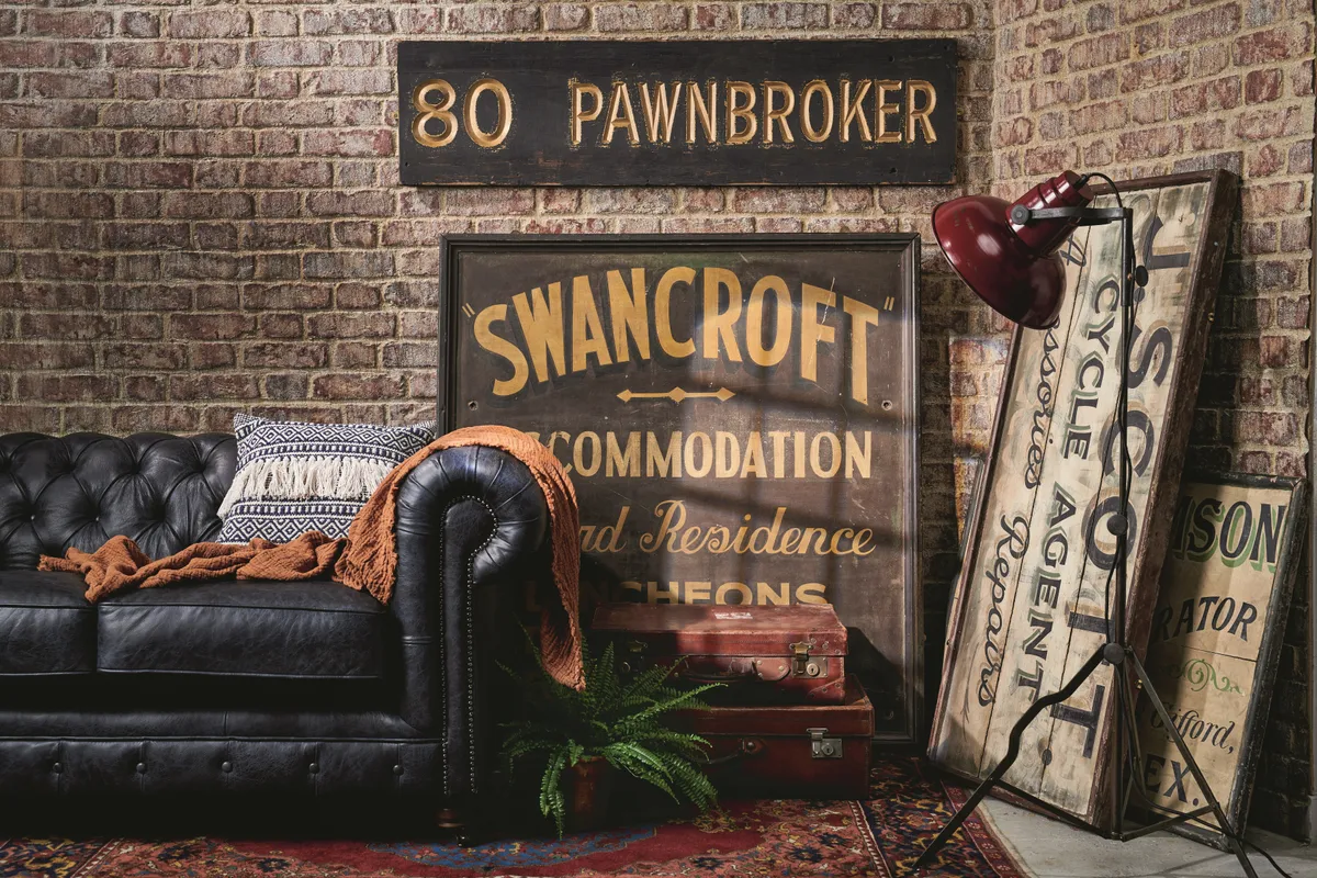An array of vintage signs leant against an exposed brick wall alongside black leather Chesterfield sofa.