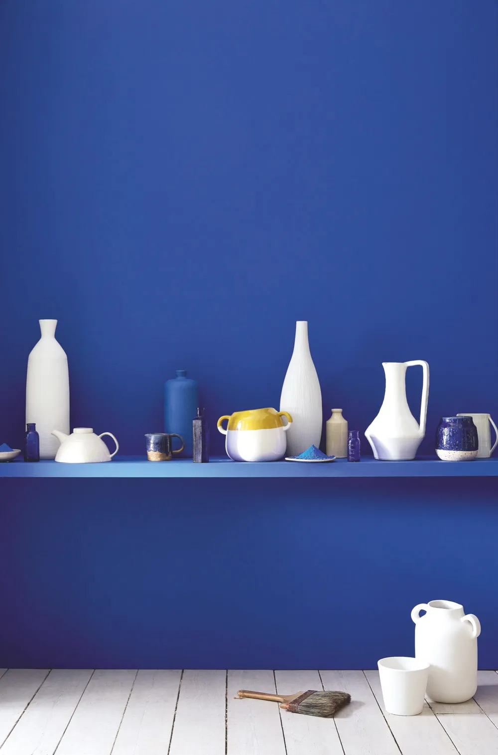 A wall painted in bright blue paint with a matching shelf. In the shelf is an array of contemporary blue, white and yellow ceramics