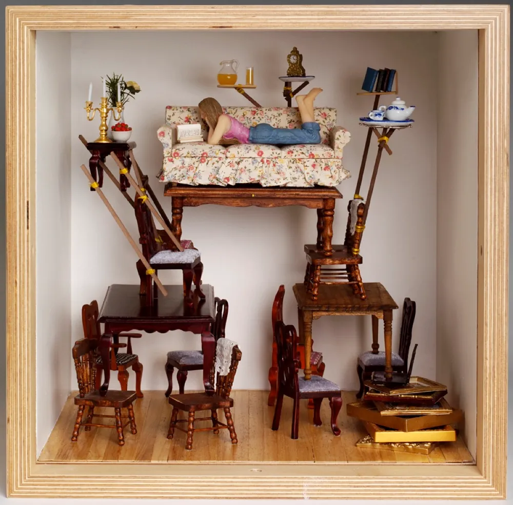 A dolls house room showing stacks of miniature antique furniture and a teenage girl lounging on a chintz sofa