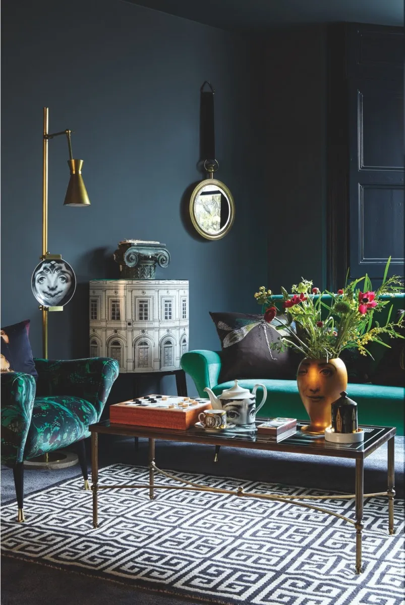 A dark grey living room featuring a green velvet sofa and armchair, plus Fornasetti plates and a Fornasetti drinks cabinet