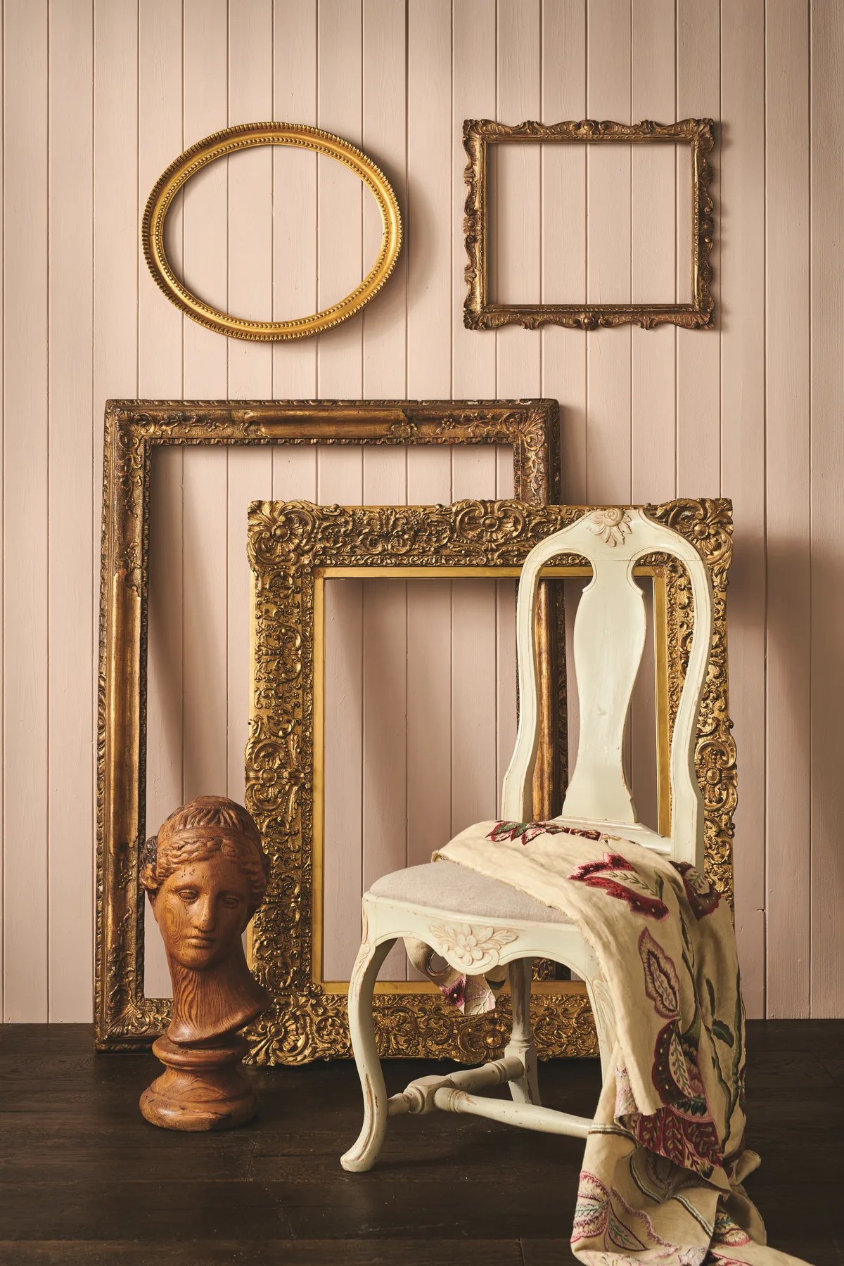 Oversized gold frames leant against a blush pink wall with a washed white chair.
