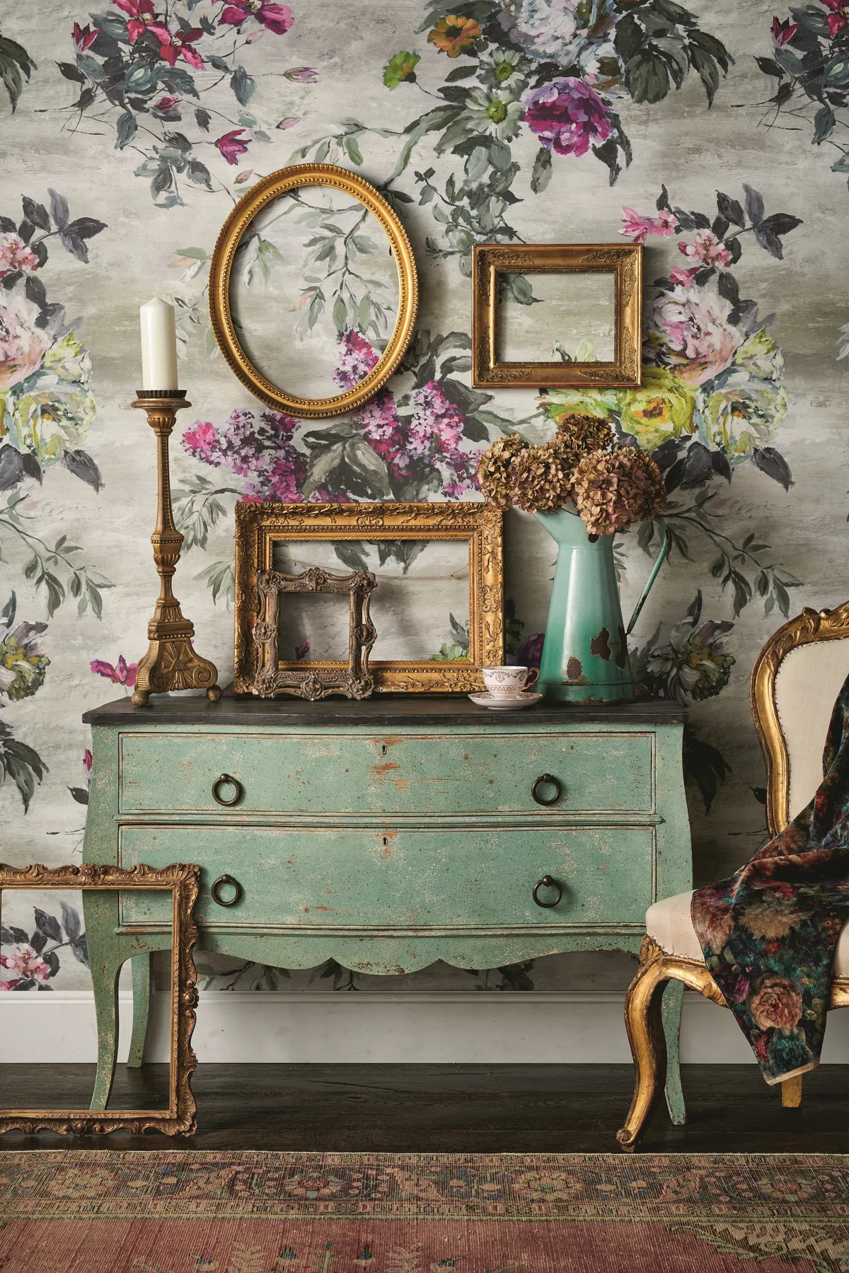 An antique commode against statement floral wallpaper, gold frames in different sizes sit atop the commode.