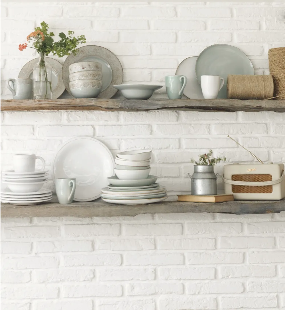 Rustic wood shelves adorned with pale-coloured plates, dishes and mugs on an exposed brick wall painted white