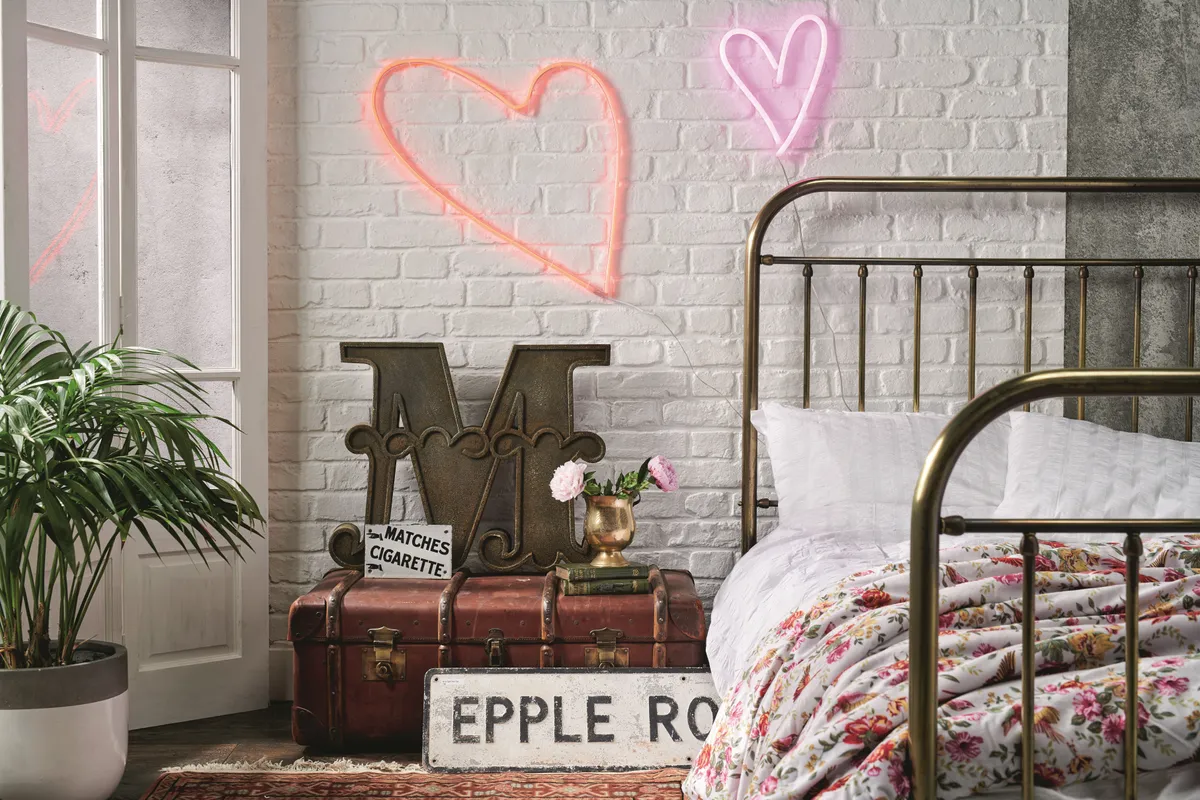 An industrial bedroom with a copper bed frame and a vintage trunk as a bedside table. There are heart-shaped neon lights on the wall.