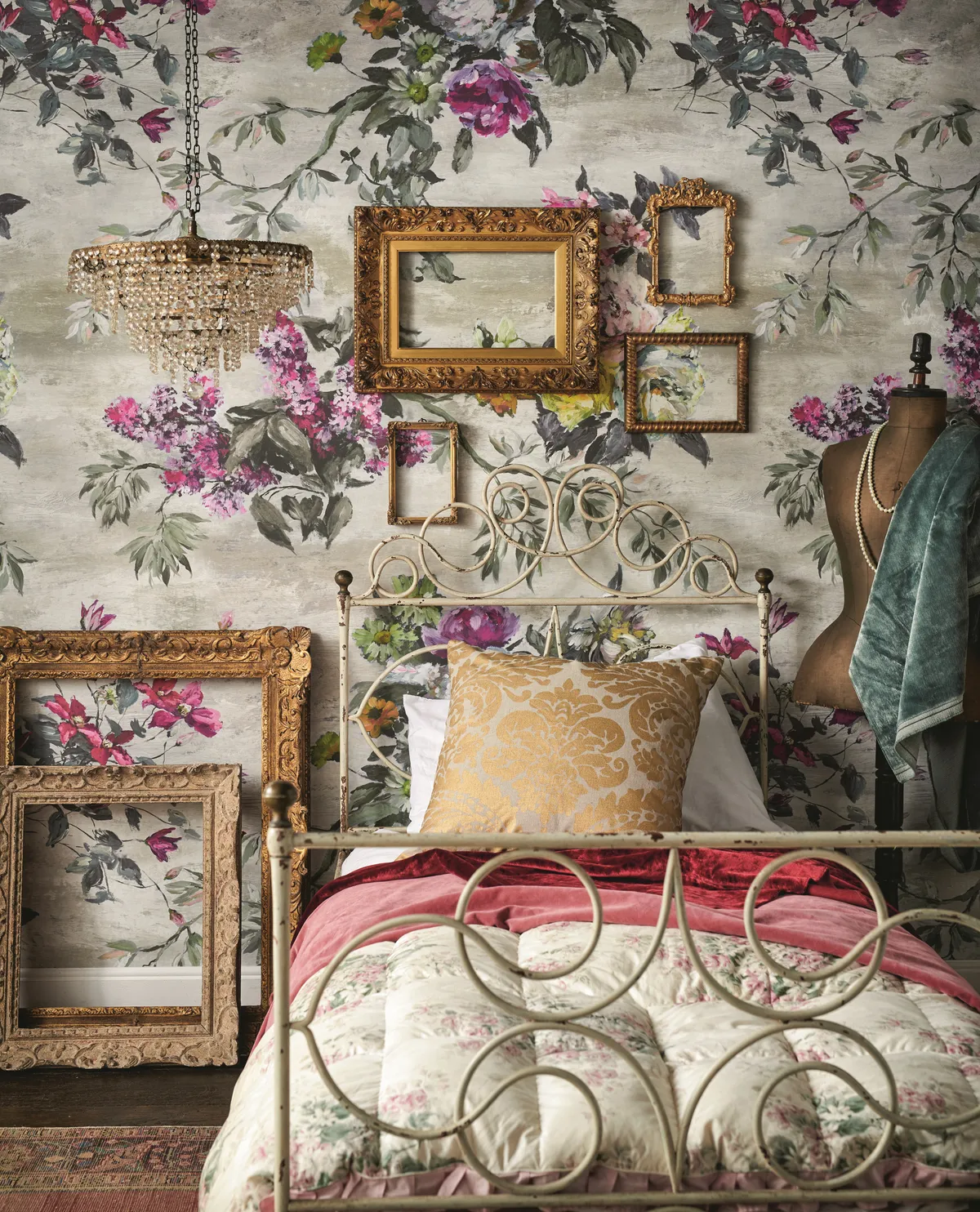 A vintage metal single bed against bold floral wallpaper. A cluster of antique frames is displayed above the headboard.