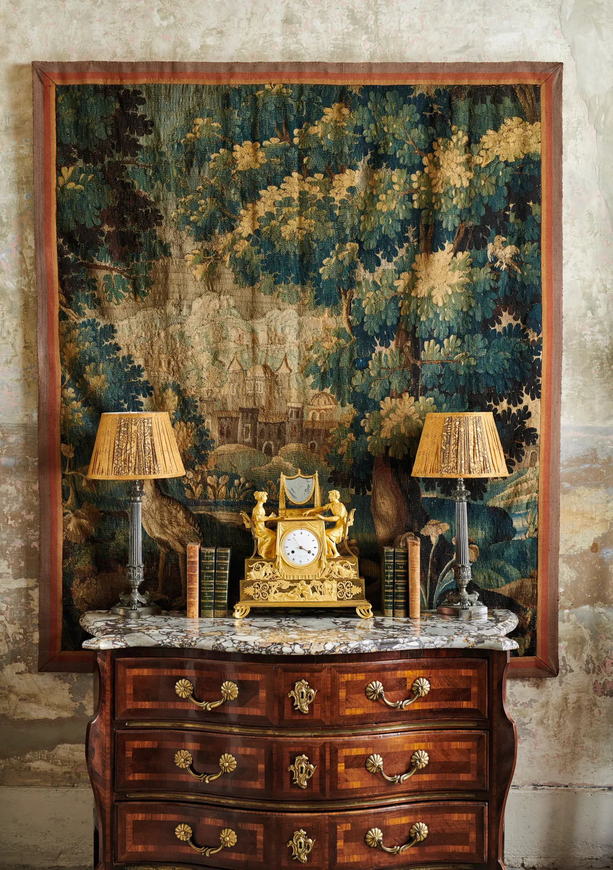 A curved antique commode decorated with an old ormolu clock flanked with table lamps. A large-scale green and brown tapestry hangs behind.
