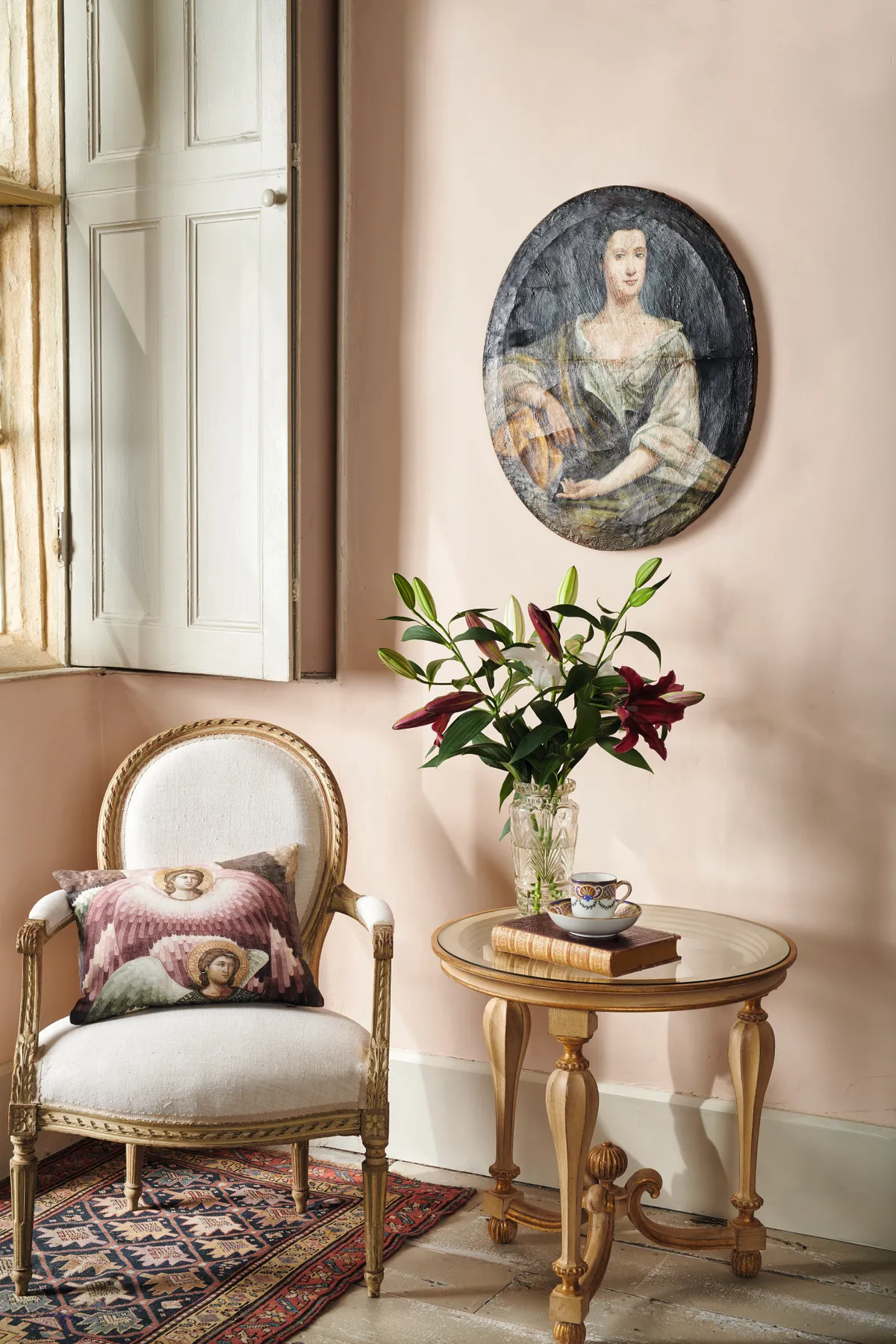 An pretty reading corner featuring a peach embroidered chair and a low table topped with a bunch of lilys and a saucer of tea.