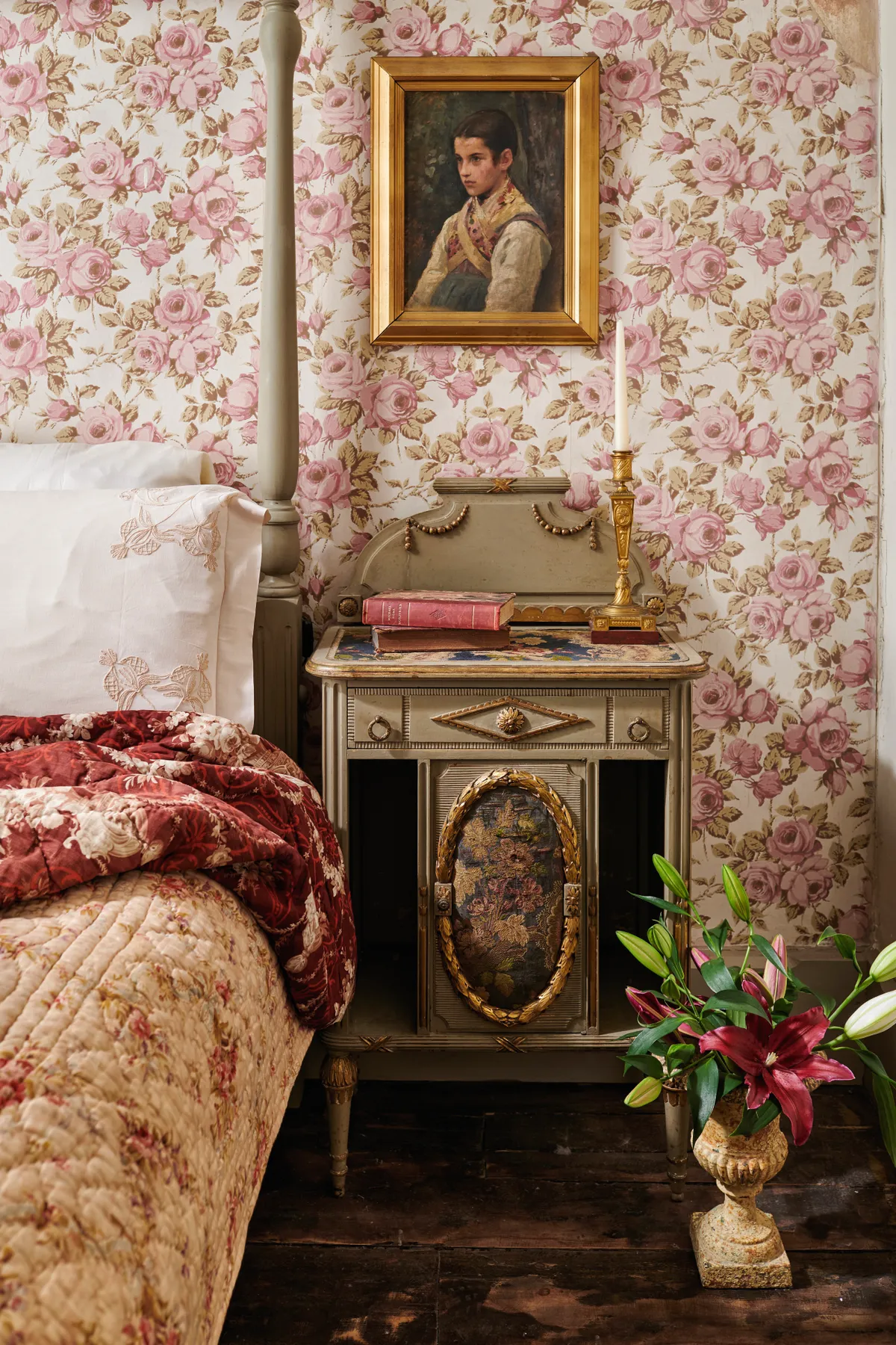 An antique four poster bed against bold floral wallpaper and a gold bedside table.