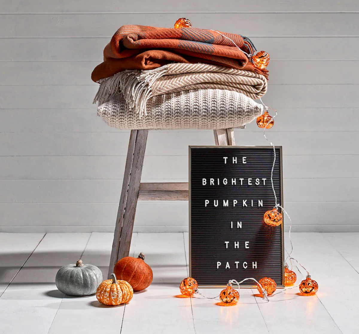 A string of illuminated pumpkin fairy lights alongside a washed wooden stool, woollen orange blankets and a vintage-style letter board.
