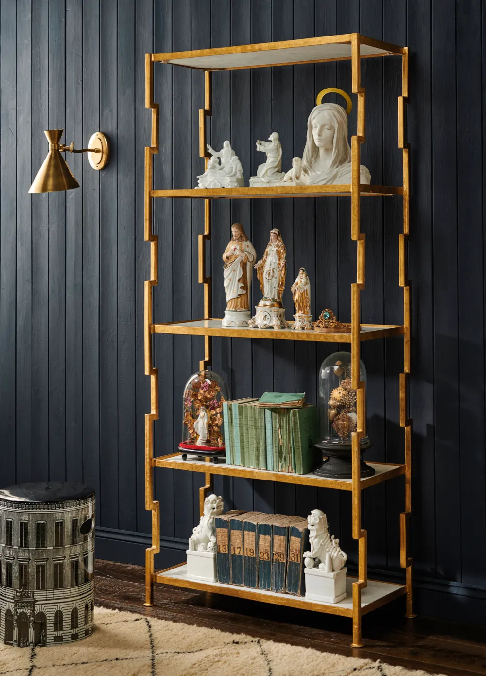 Antique and vintage figures displayed on a gold shelf next to a printed stool with antique books, brass wall light and display domes