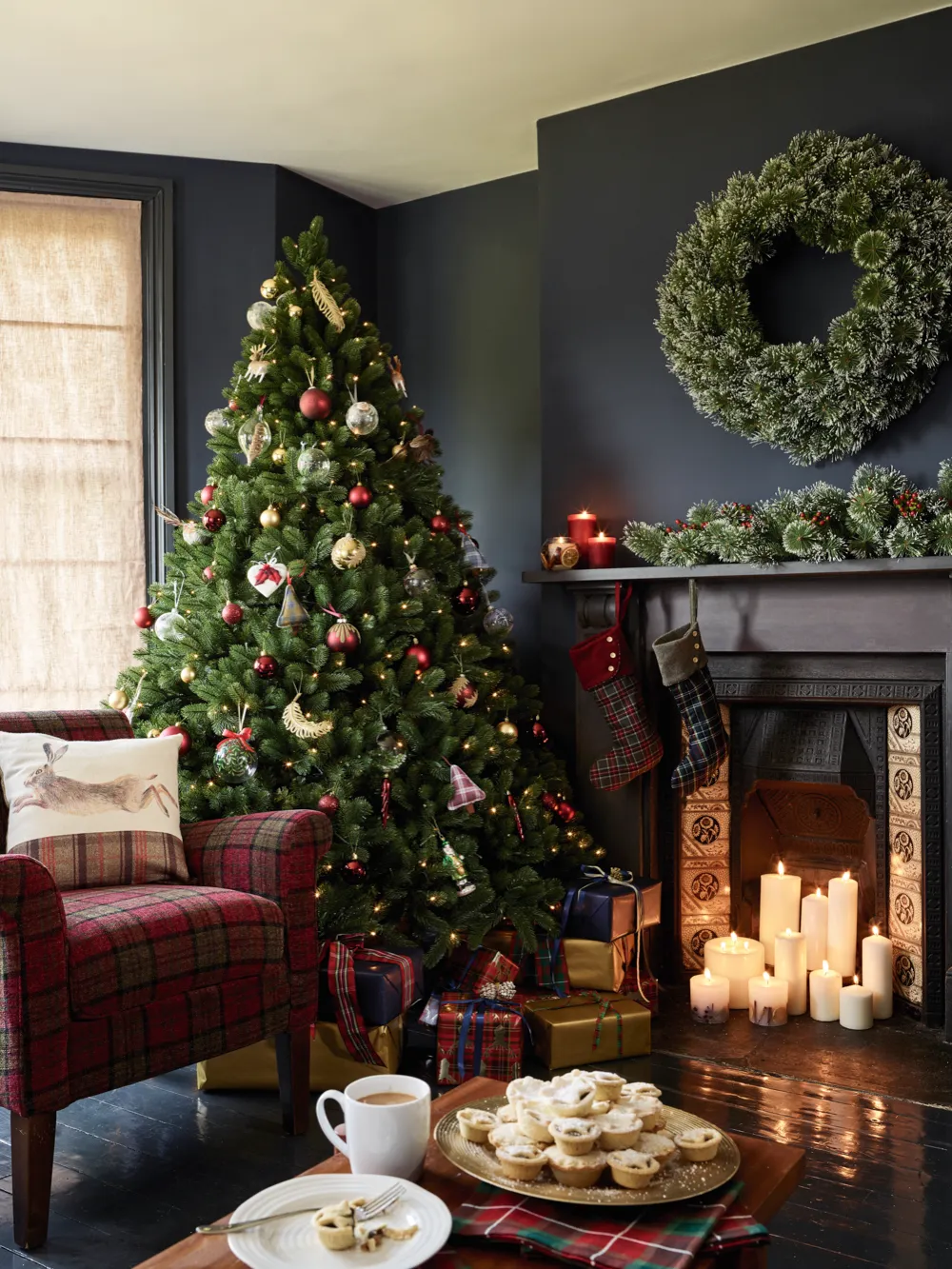 A large green Christmas tree stands next to a fireplace lit with pillar candles. Red and gold decorations fill the tree. Layered with traditional tartan this classic look oozes comfort and class