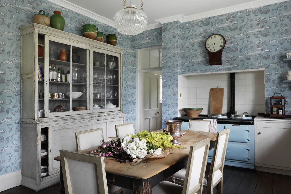 A homely VSP Interiors Devon kitchen filled with an antique wall clock, rustic table, French confit pots and a pale blue Aga
