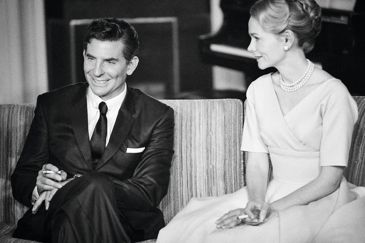 A black and white film still from Netflix's Maestro featuring Bradley Cooper and Carey Mulligan sitting on a sofa