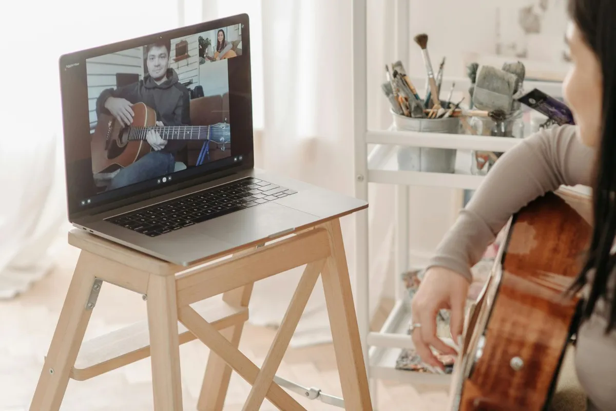 A woman in a guitar lesson with the teacher on a screen on a laptop sat on top of a stool
