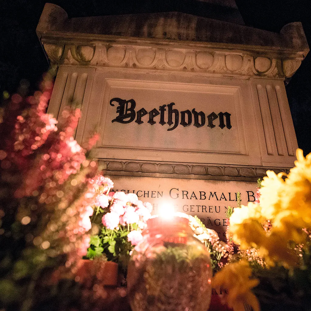 A close-up image of a grave with the word 'Beethoven' written on it, candles lit and flowers surrounding it