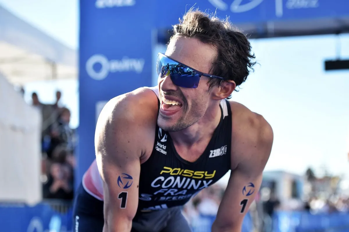 French triathlete Dorian Coninx wearing sunglasses and smiling to someone off camera puts his hands on his knees as he catches his breath after winning the 2022 Bergen World Cup
