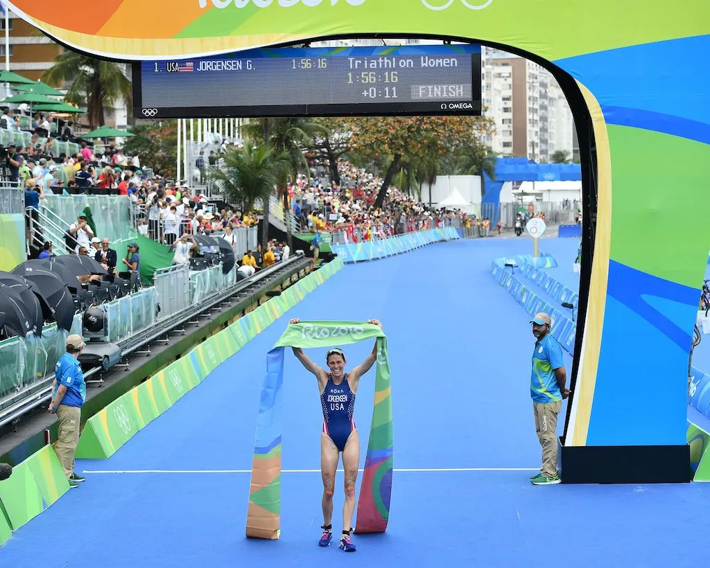 USA's Gwen Jorgensen crosses the line at the 2016 Rio Olympic Game to win gold, holding the finisher's tape aloft, with crowds and media on the left hand of the image
