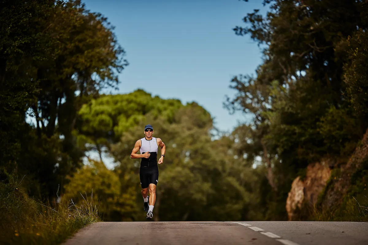 Man running over the crest of a small hill on the road, wearing a tri-suit, cap and glasses