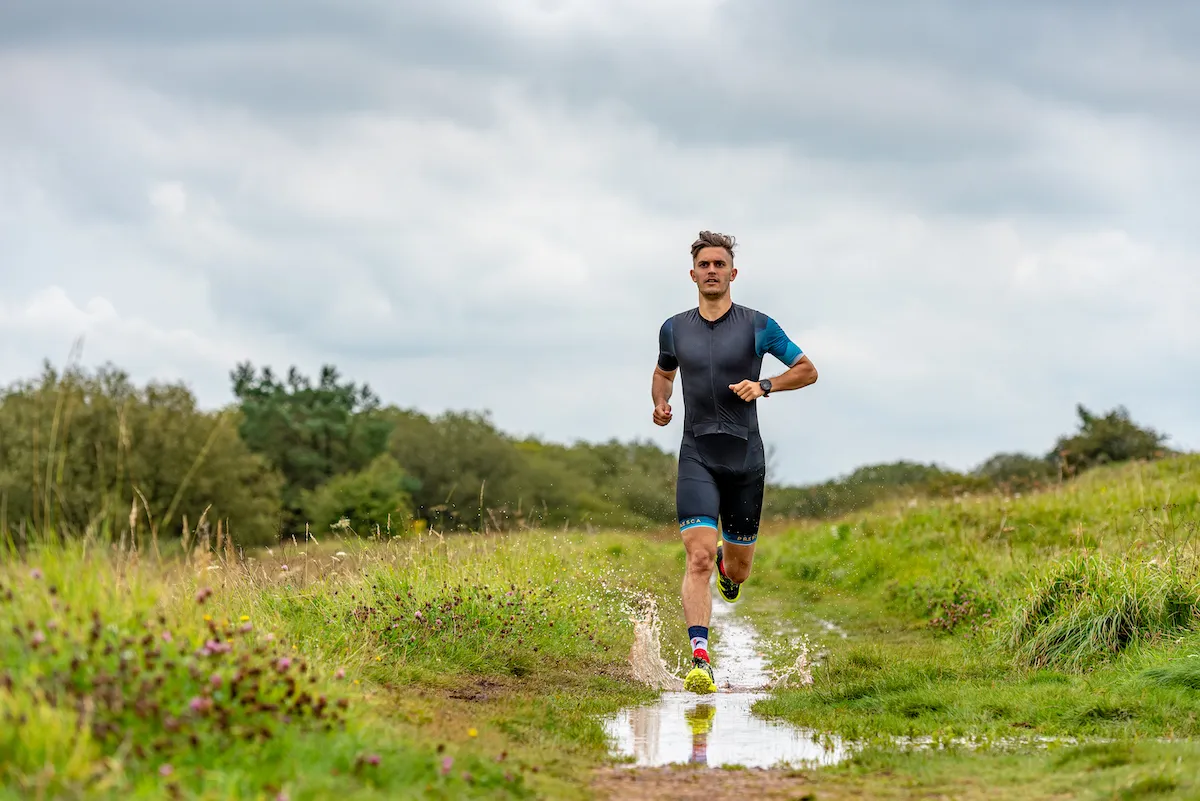 Male triathlete wearing a tri-suit running through a puddle on a path through a field