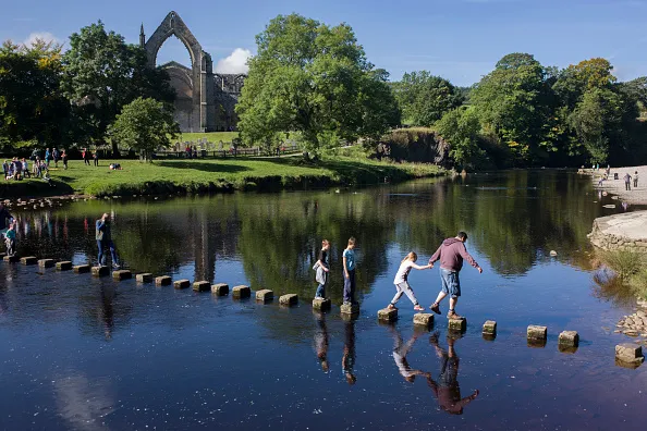 Families risk falling in the River Wharfedale while walking over the stepping stones at Augustinian Bolton Priory, North Yorkshire. Carefully stepping stone by stone, a young girl holds the hand of an adult who guides her across to the other side. The monastery was founded in 1154 by the Augustinian order, on the banks of the River Wharfe. The land at Bolton, as well as other resources, were given to the order by Lady Alice de Romille of Skipton Castle in 1154. It is now a popular loaction for families and walkers who can trek the River Wharfe upstream into ancient woodland.