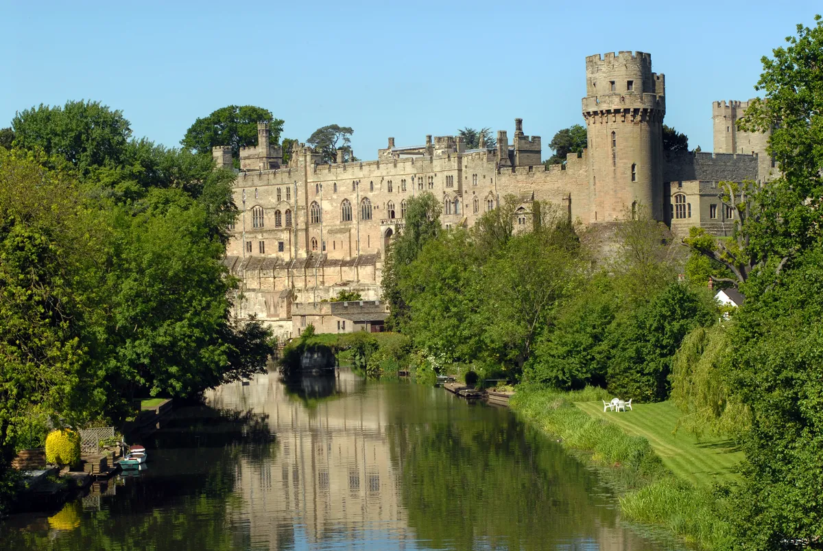 Warwick Castle surrounded by trees with a river in the foreground