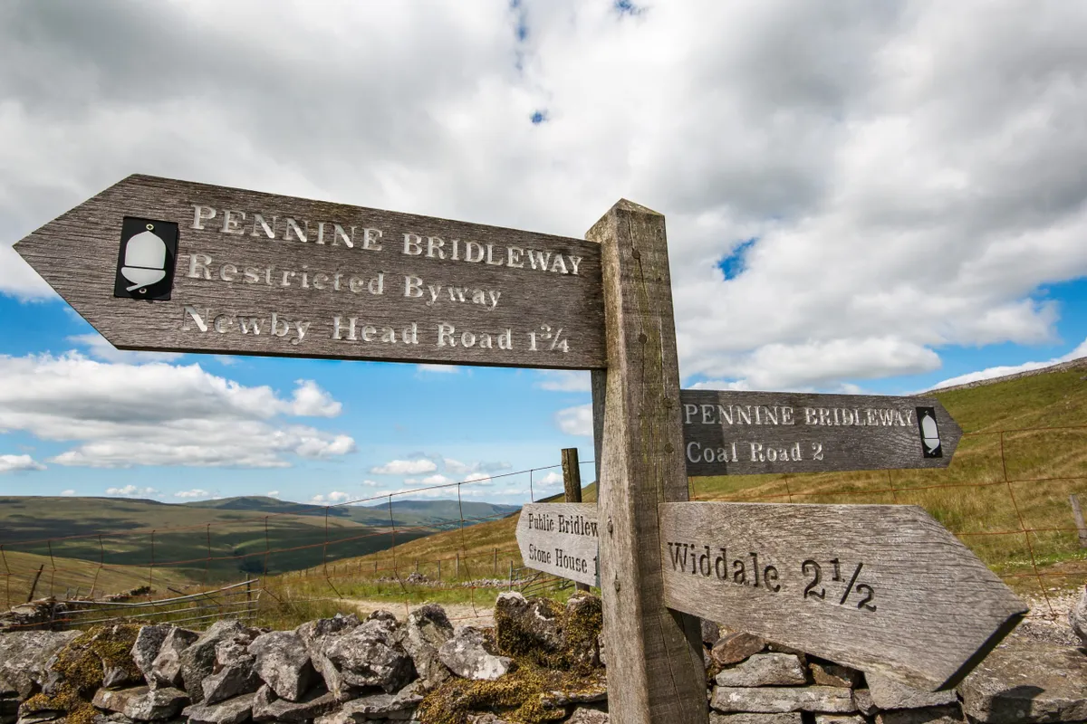 Fingerpost or signpost of walking distances at crossroad of paths and Pennine Bridleway with dry stone wall and open moorland hillsides in background