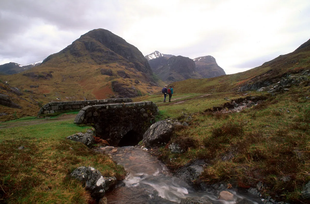 A Couple Walk Beyond An Old Stone Bridge Whilst Hillwalking In Glen Coe, With A View To Mountains Behind, VisitScotland