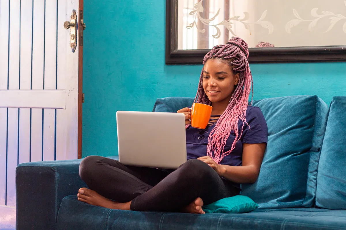 Young black woman with pink braids using notebook sitting on blue sofa in home room