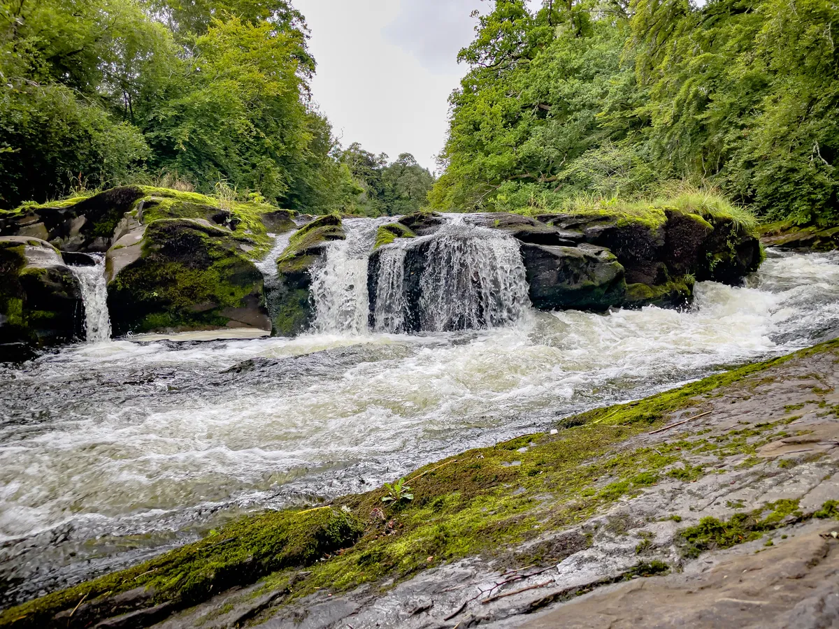 Low lying waterfalls of Allan Water in perthshire with green trees behind and grey sky