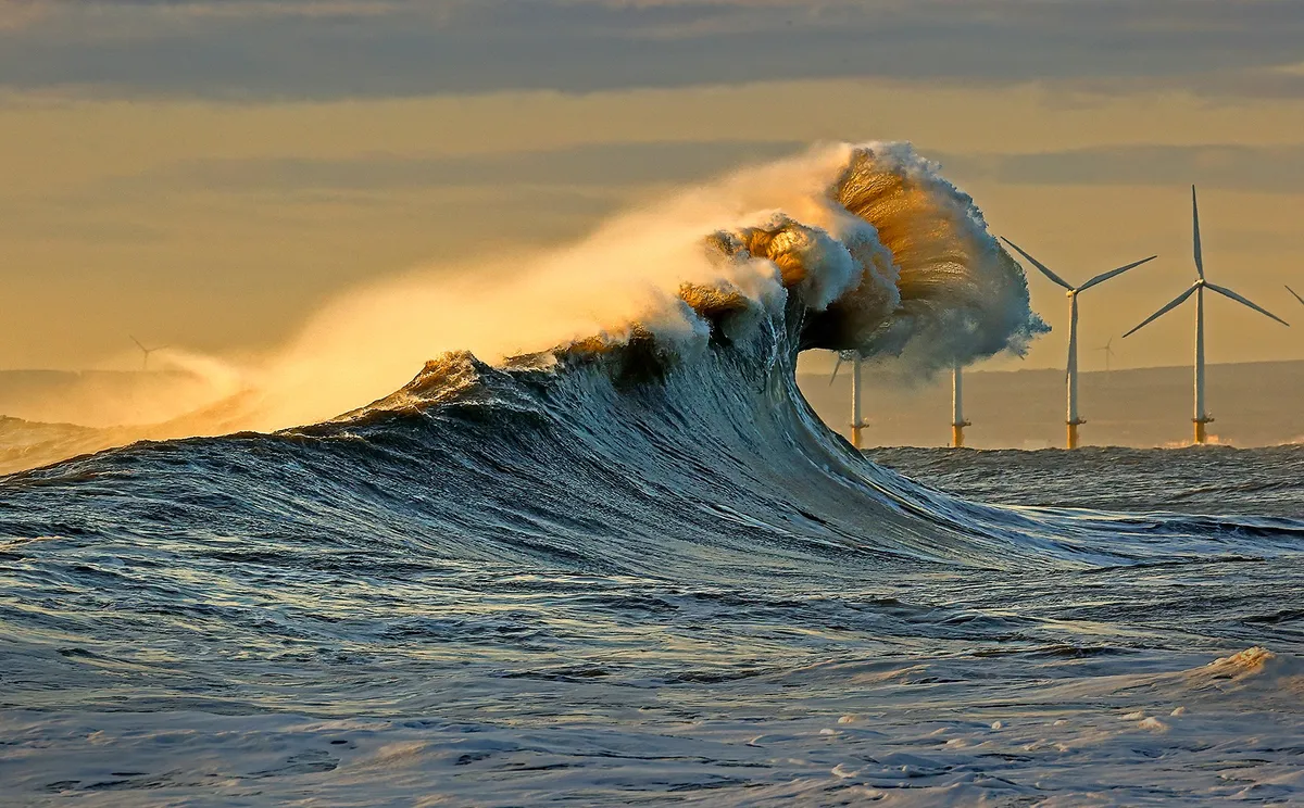Waves curl over dramatically on the sea with windmills in the background
