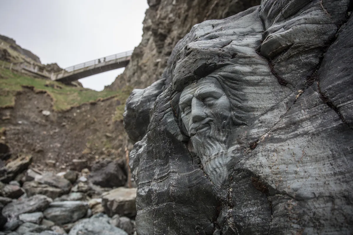 TINTAGEL, UNITED KINGDOM - APRIL 28: Visitors walk on a bridge above a carving of Merlin at Tintagel Castle in Tintagel on April 28, 2016 in Cornwall, England. The English Heritage managed site and the nearby town have long been associated with the legend of King Arthur and continue to attract large visitor numbers. However, efforts by English Heritage to update the visitor experience with the Gallos sculpture, along with a rock carving of Merlin's face, which English Heritage say are inspired by the legend of King Arthur and Tintagel Castles royal past, have met with criticism from some Cornish nationalists and historians. (Photo by Matt Cardy/Getty Images)