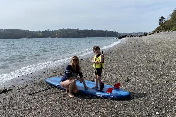 Maria Hodson on the shingle of Grebe beach with a stand up paddle and her son wearing a life jacket ready to launch at the shoreline