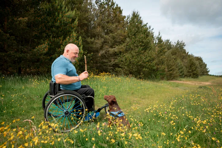A mature man with paraplegia, using his wheelchair in a nature reserve on a sunny summers day. He is wearing casual clothing and is throwing a stick for his dog, a brown Patterdale Terrier.