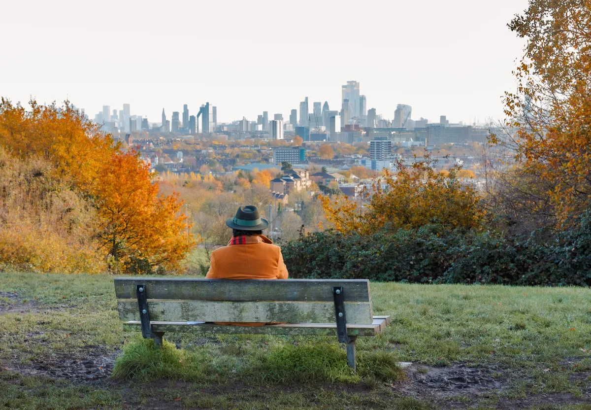 Senior man sitting on park bench looking at London city skyline from Hampstead Heath