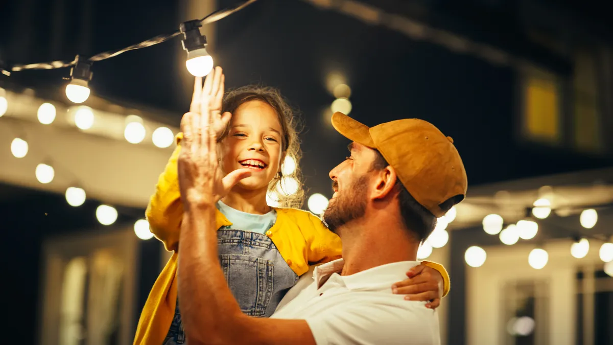 Father and the  daughter he is carrying high-five under glowing string lights