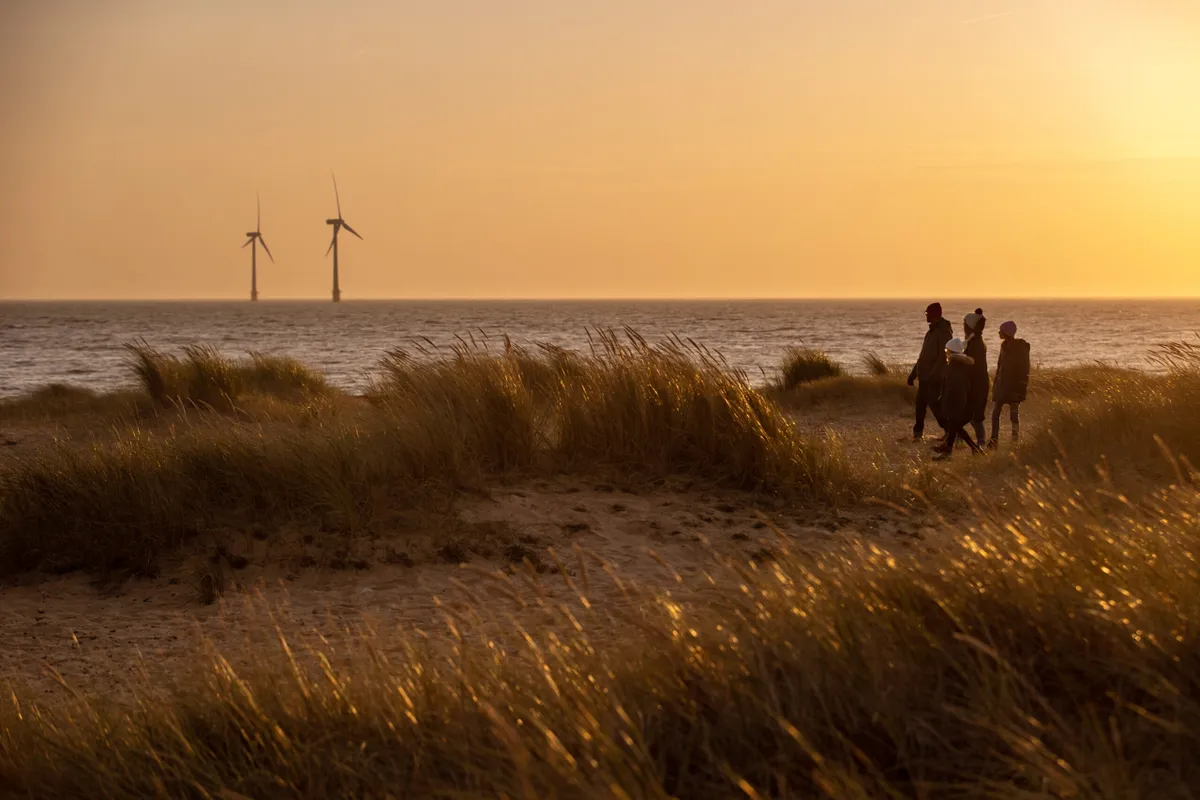Family of four walking in in sand dunes at golden hour, two wind turbines out at see in the distance