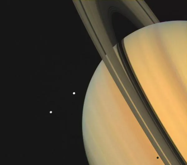 Saturn and two of its moons photographed in 1980 by Voyager 1. The moons, Tethys (closest to the planet) and Dione, are visible as bright spots in space next to the gas giant. Tethys's shadow can also be seen on Saturn itself. (Credit: NASA/JPL)