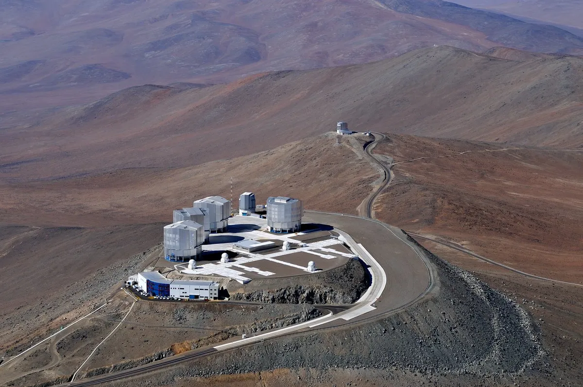 A bird soaring over the remote, sparsely populated Atacama Desert in northern Chile — possibly the driest desert in the world — might be surprised to come upon the technological oasis of ESO’s Very Large Telescope (VLT) at Paranal. The world’s most advanced ground-based facility for astronomy, the site hosts four 8.2-metre Unit Telescopes, four 1.8-metre Auxiliary Telescopes, the VLT Survey Telescope (VST), and the 4.1-metre Visible and Infrared Survey Telescope for Astronomy (VISTA), seen in the distance on the next mountain peak over from the main platform. This aerial view also shows other structures, including the Observatory Control Room building, on the main platform’s front edge.
