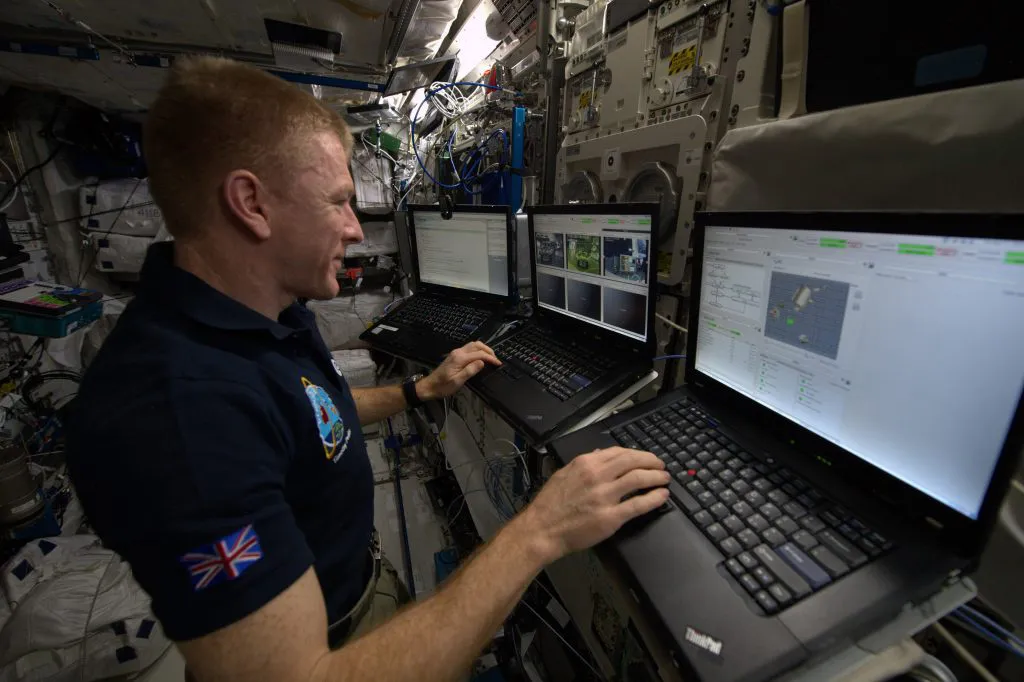 Tim Peake at a bank of laptops during his space mission, a UK flag on his arm