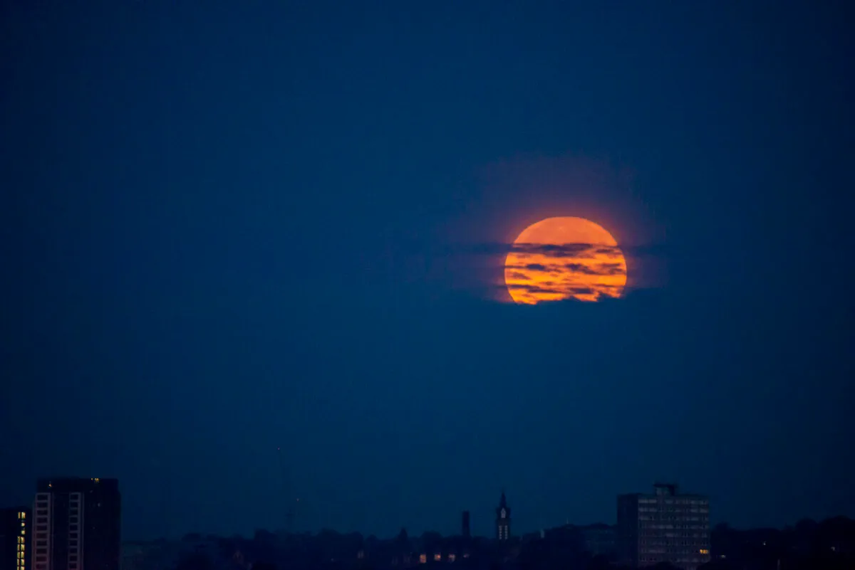 An image of a glowing orange full Moon rising through the clouds.