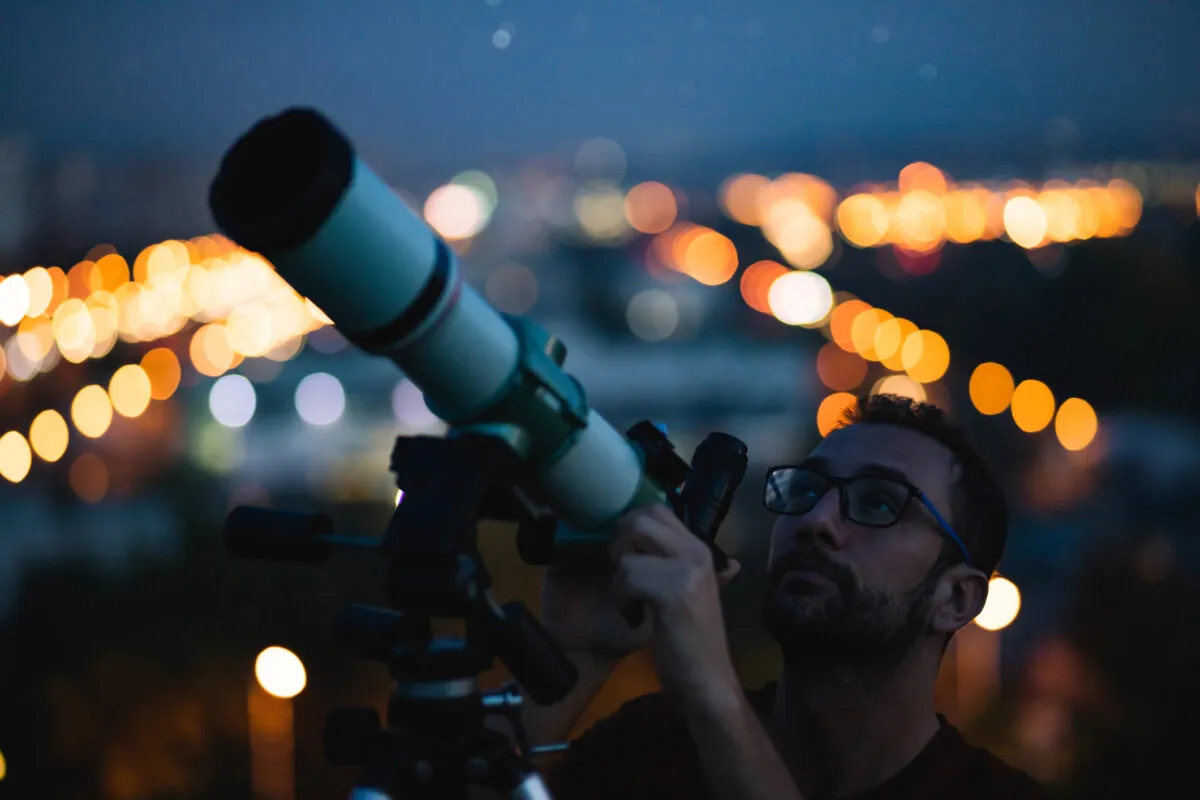 Man observing the night sky through a telescope with blurred city lights in the background