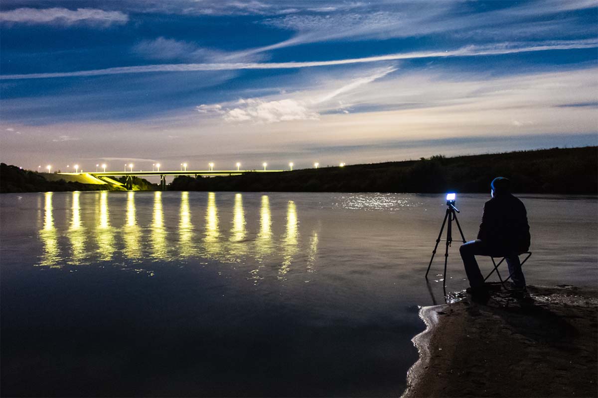 Photo of a man standing in front of a smartphone on a tripod, capturing a nighttime landscape