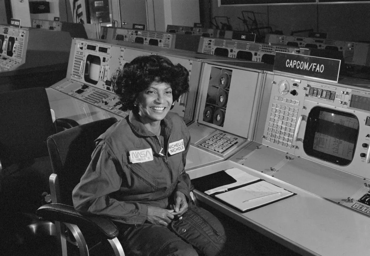 Nichelle Nichols in a NASA jumpsuit in front of a control panel.