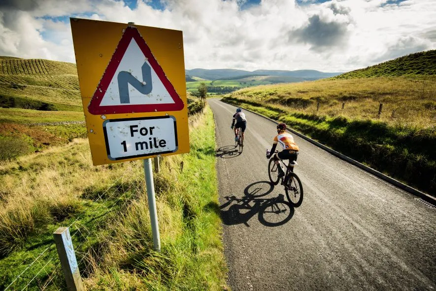 Cyclists ride pass a road sign warning of sharp bends