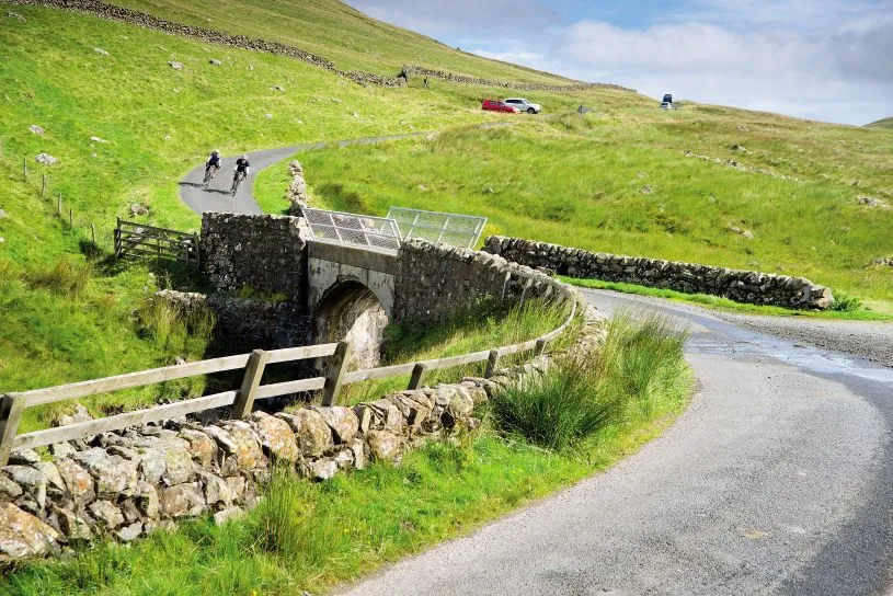 Two cyclists descend a hill towards a stone bridge in the Scottish Borders